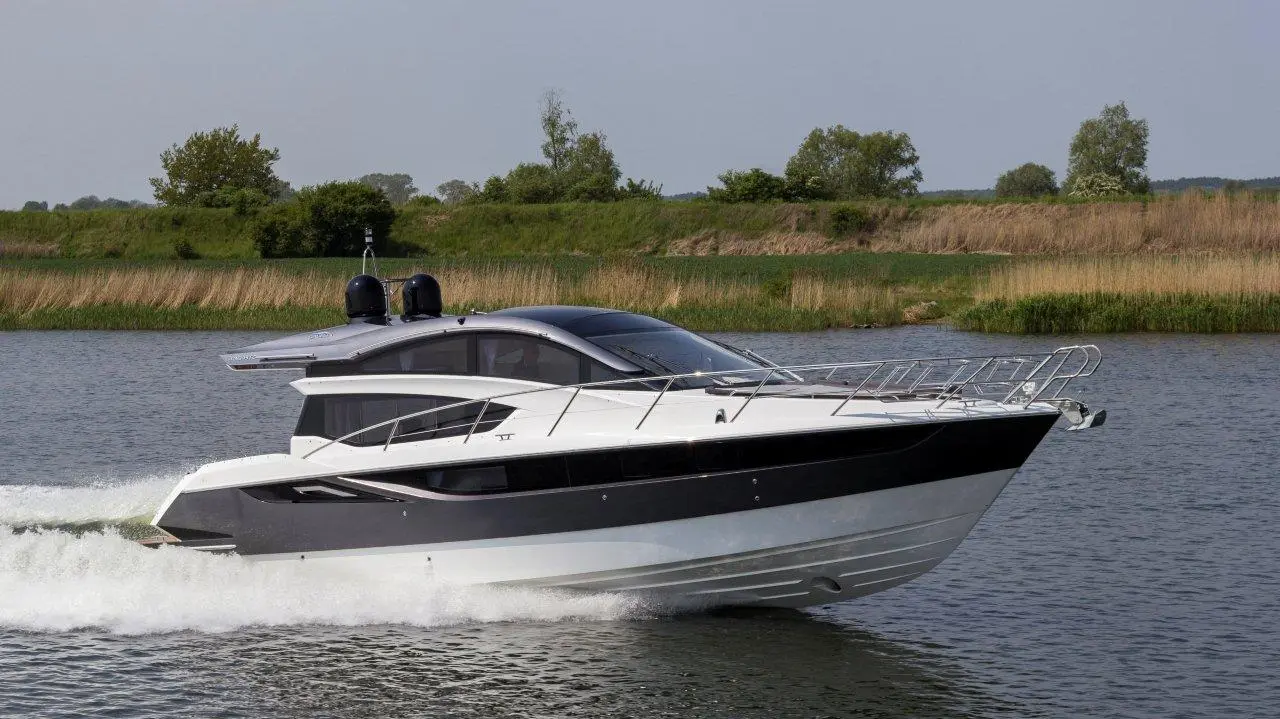 A sleek motorboat with a black and white design speeds across a calm lake surrounded by lush greenery and reeds.