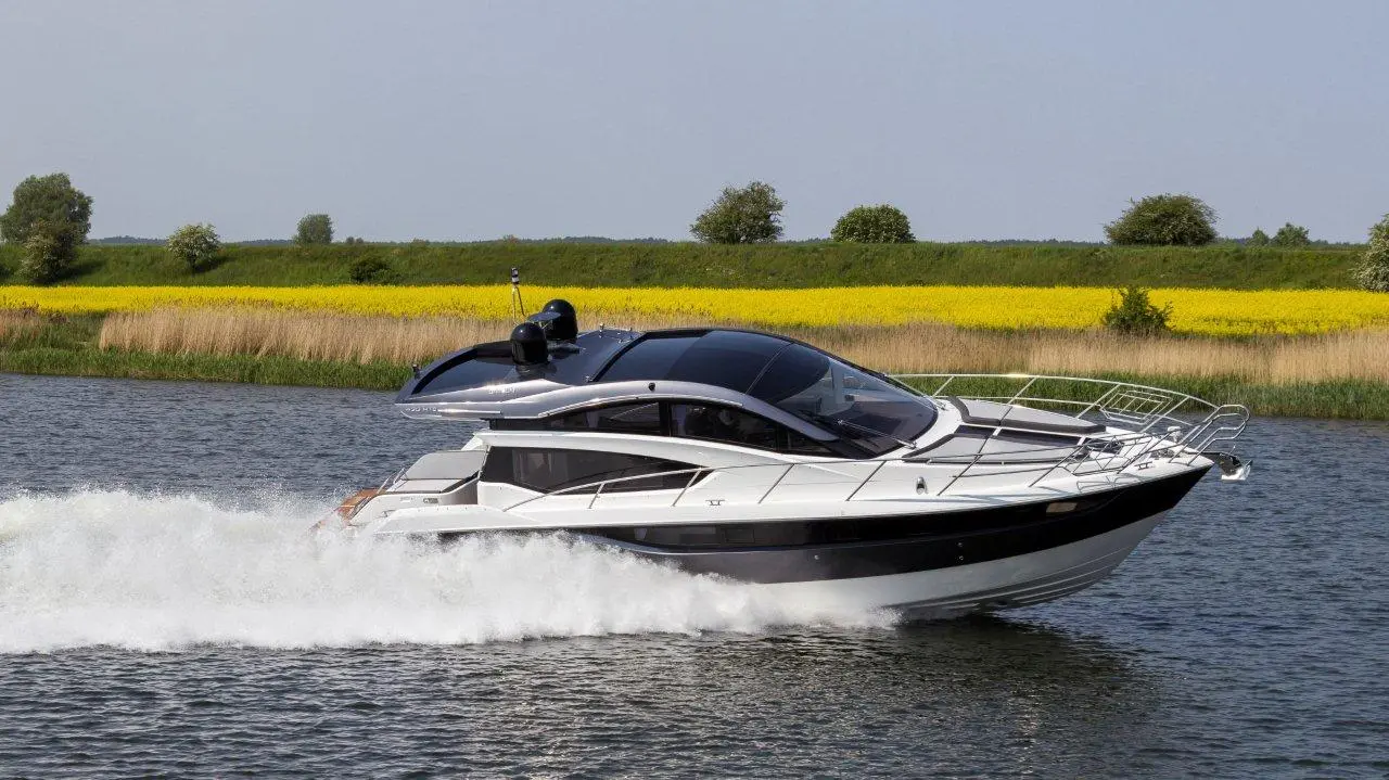 A sleek motorboat cuts through calm water, surrounded by lush greenery and fields of vibrant yellow flowers in the background.