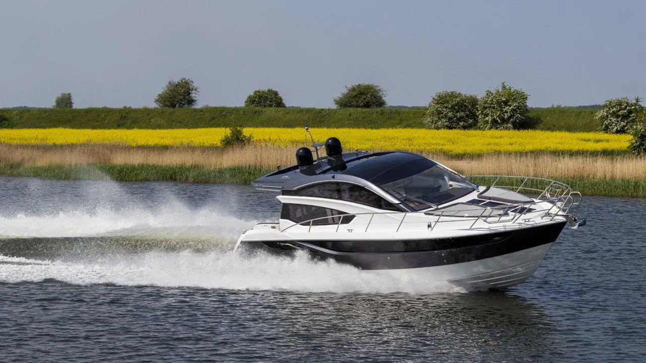 A modern motorboat speeds through calm waters, with vibrant yellow fields and green trees in the background.
