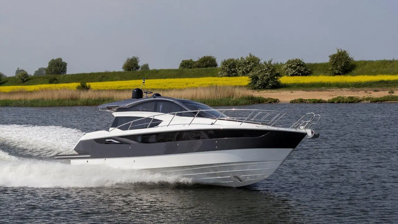 A sleek motor yacht speeds across a calm river, surrounded by greenery and bright yellow flowers in the background.