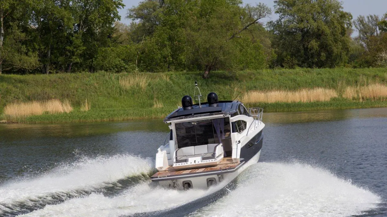 A motorboat speeds through a calm river, creating a large wake against a lush green shoreline.
