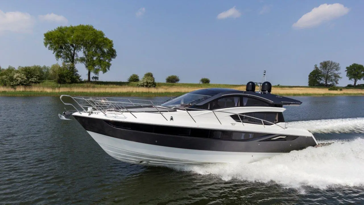 A sleek motorboat glides through calm water under a blue sky, surrounded by lush greenery and distant trees.