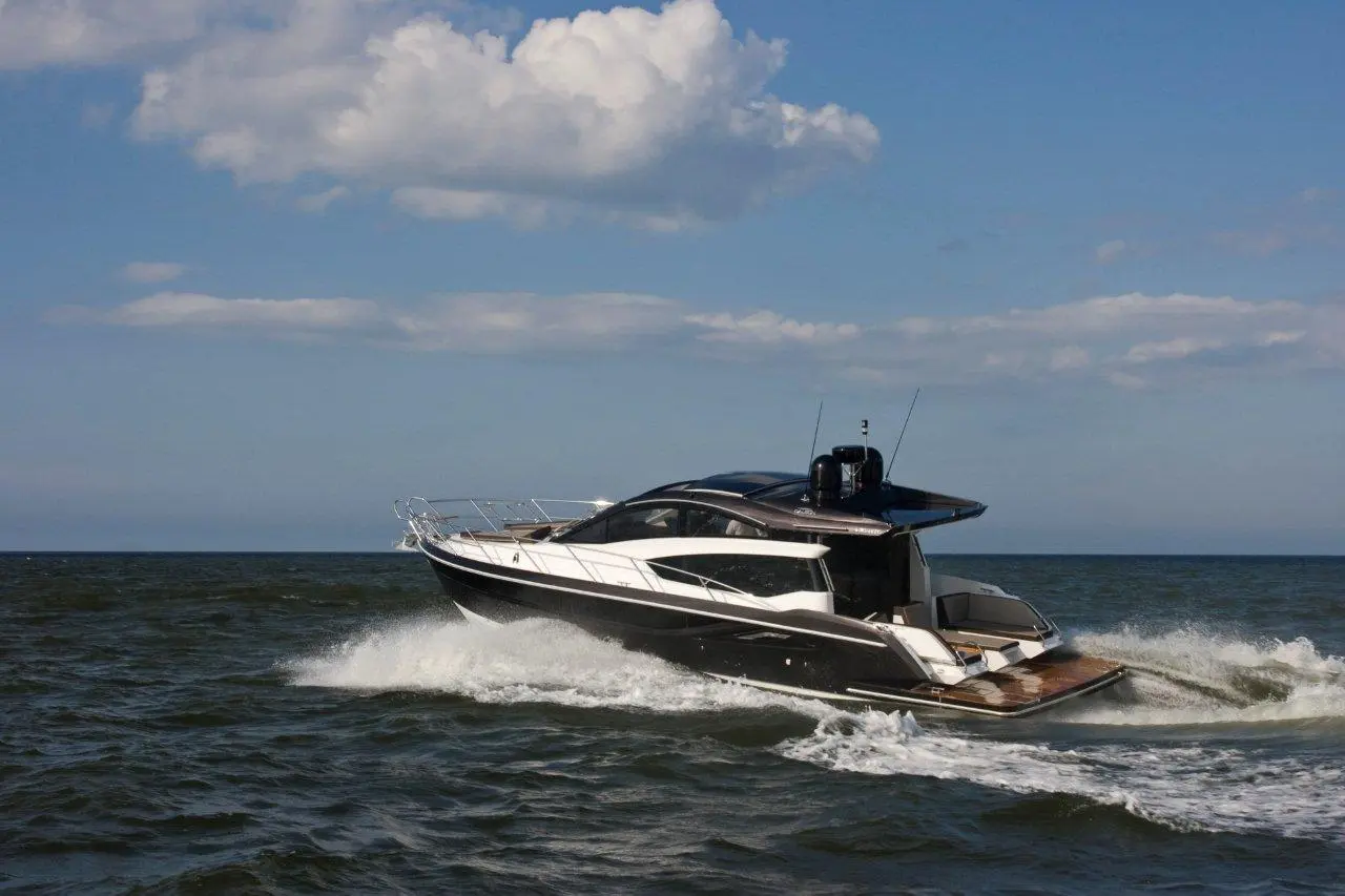 A sleek black and white motor yacht speeds across the sea, creating waves under a partly cloudy sky.