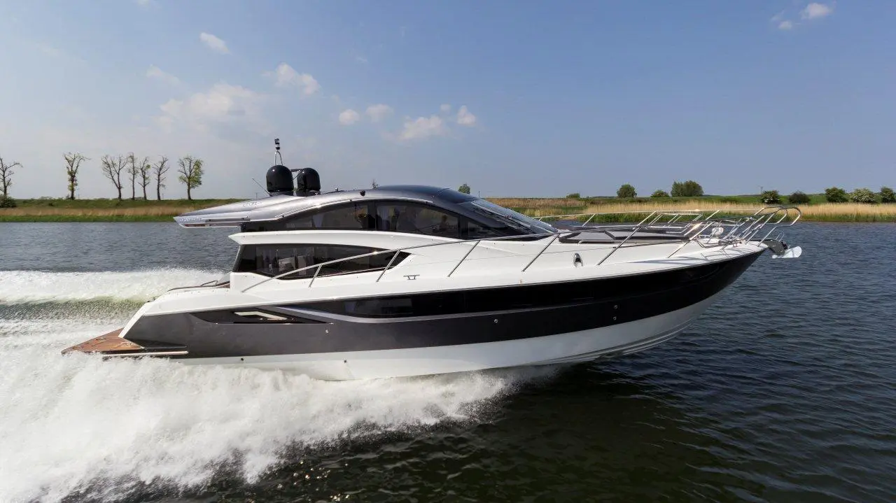 A modern yacht cruising on a river, creating waves, with trees lining the shore under a clear blue sky.
