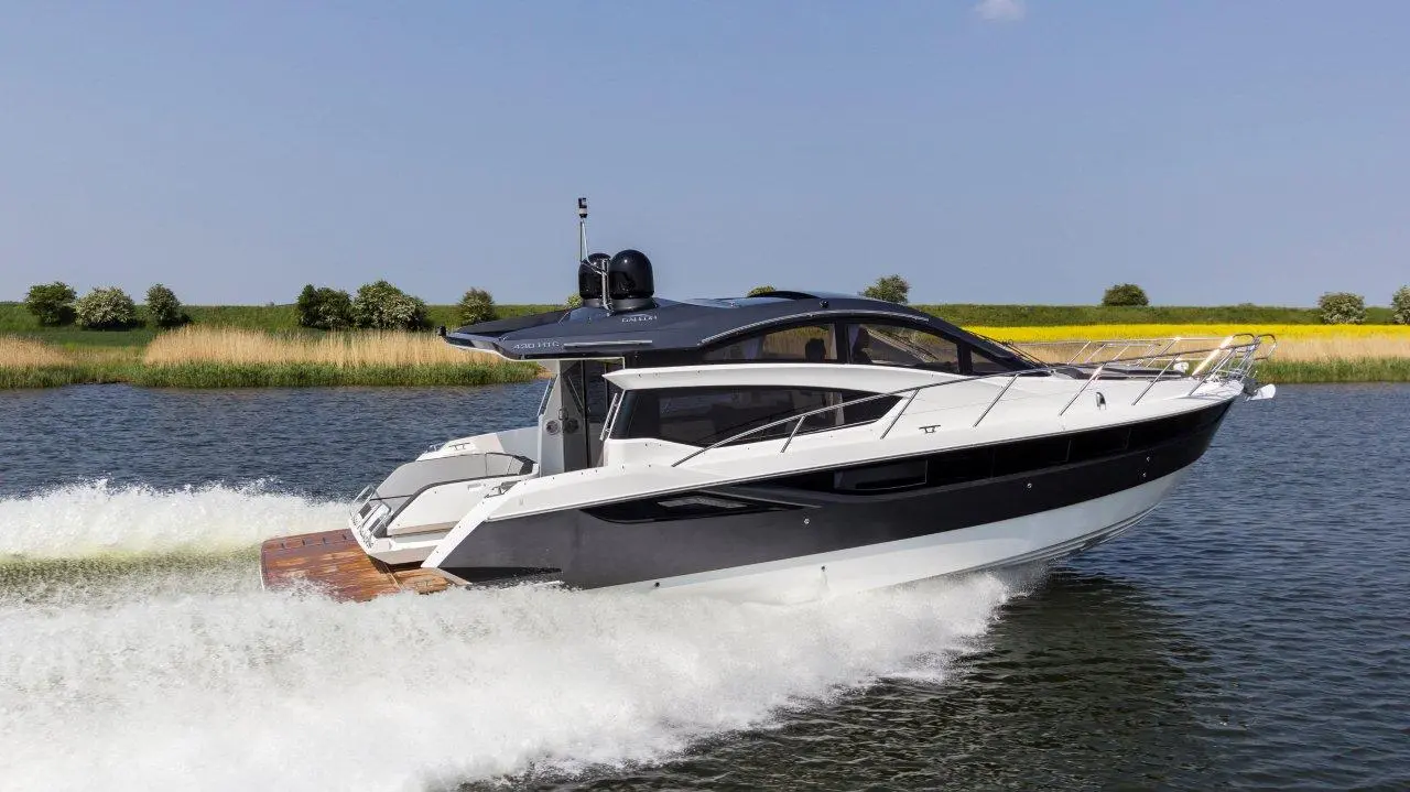 A sleek motorboat cruising on a calm river, leaving a white wake, surrounded by greenery and fields under clear blue skies.