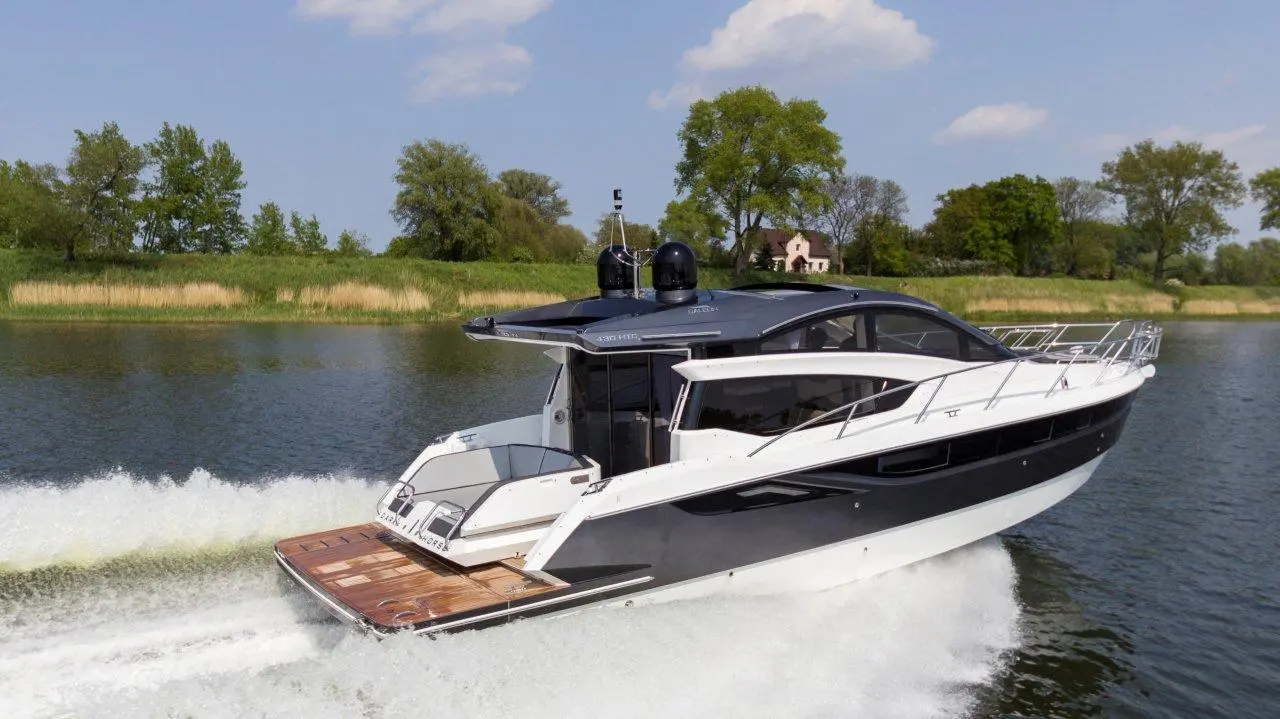 A sleek motorboat glides over water, creating wakes, with green trees and a house visible in the background under a blue sky.