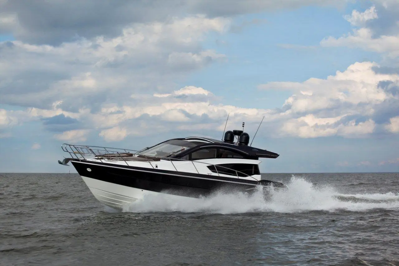 A sleek black and white motorboat speeds across the water, creating splashes against a backdrop of cloudy blue sky.