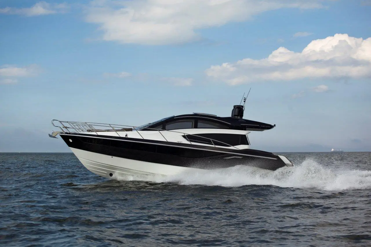 A sleek black and white motor yacht speeds over the water with waves splashing, against a blue sky with clouds.