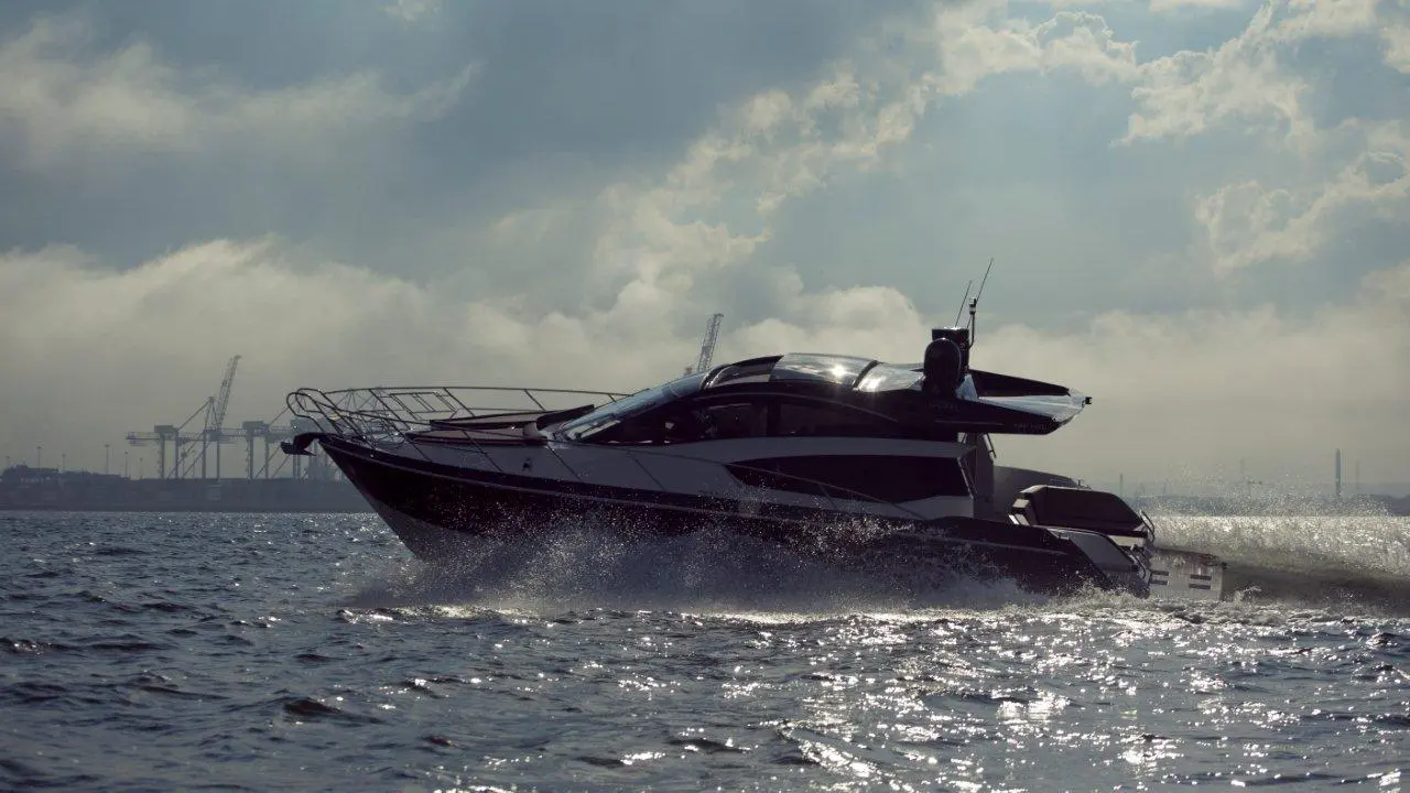 A sleek black yacht navigates through water, creating a splash against a backdrop of industrial cranes and cloudy skies.