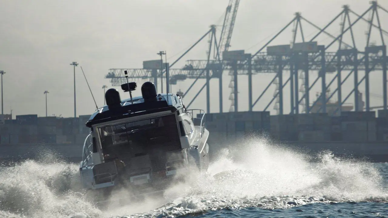 A motorboat accelerates through water with splashes, against a backdrop of shipping cranes and containers in a port.