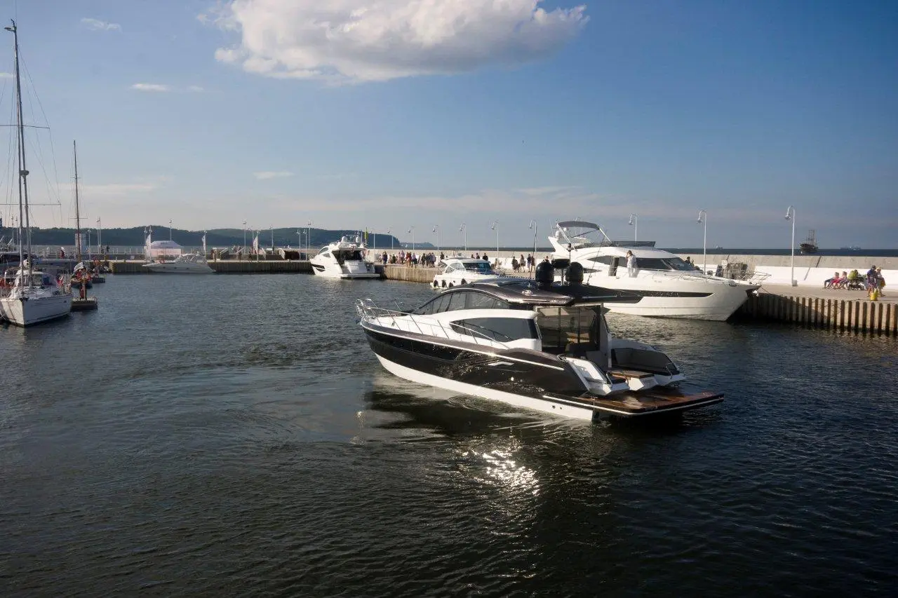 Marina scene with various boats, including a black and white yacht, docked by a lively waterfront with people.