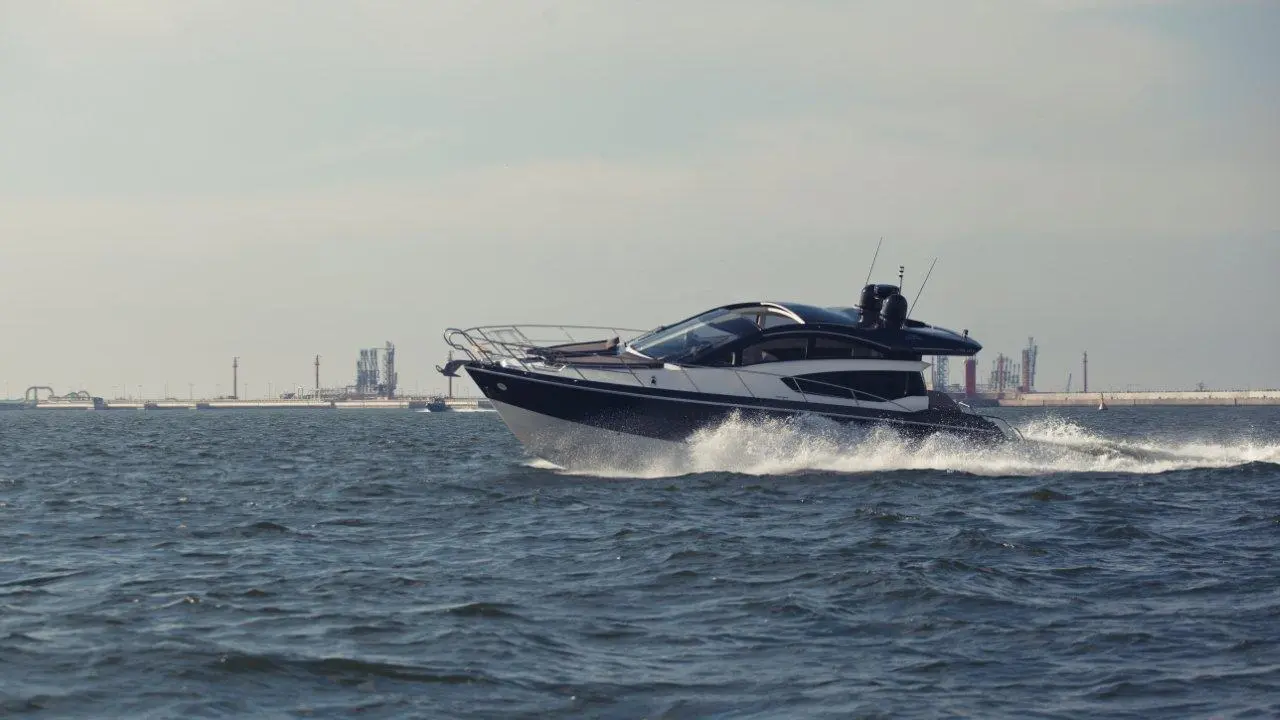A sleek motorboat speeds across water, leaving a wake, with industrial structures visible in the distant background.