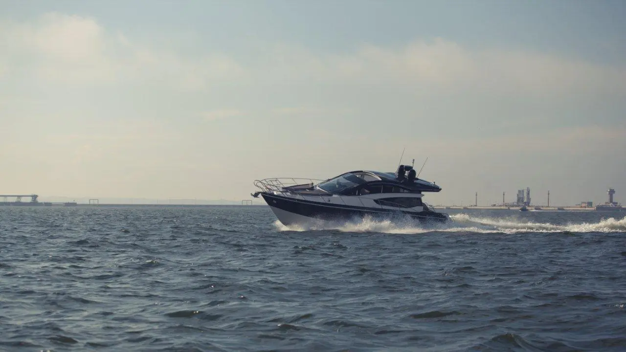 A sleek motorboat cruises across a calm sea, with industrial structures visible on the horizon.
