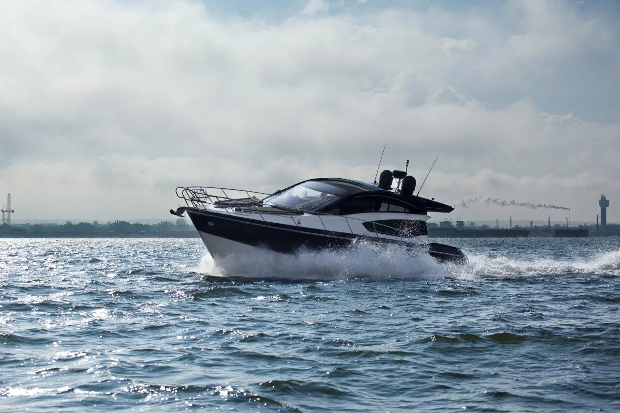A sleek black and white motorboat speeds across the water, with a cloudy sky and distant industrial landscape in the background.