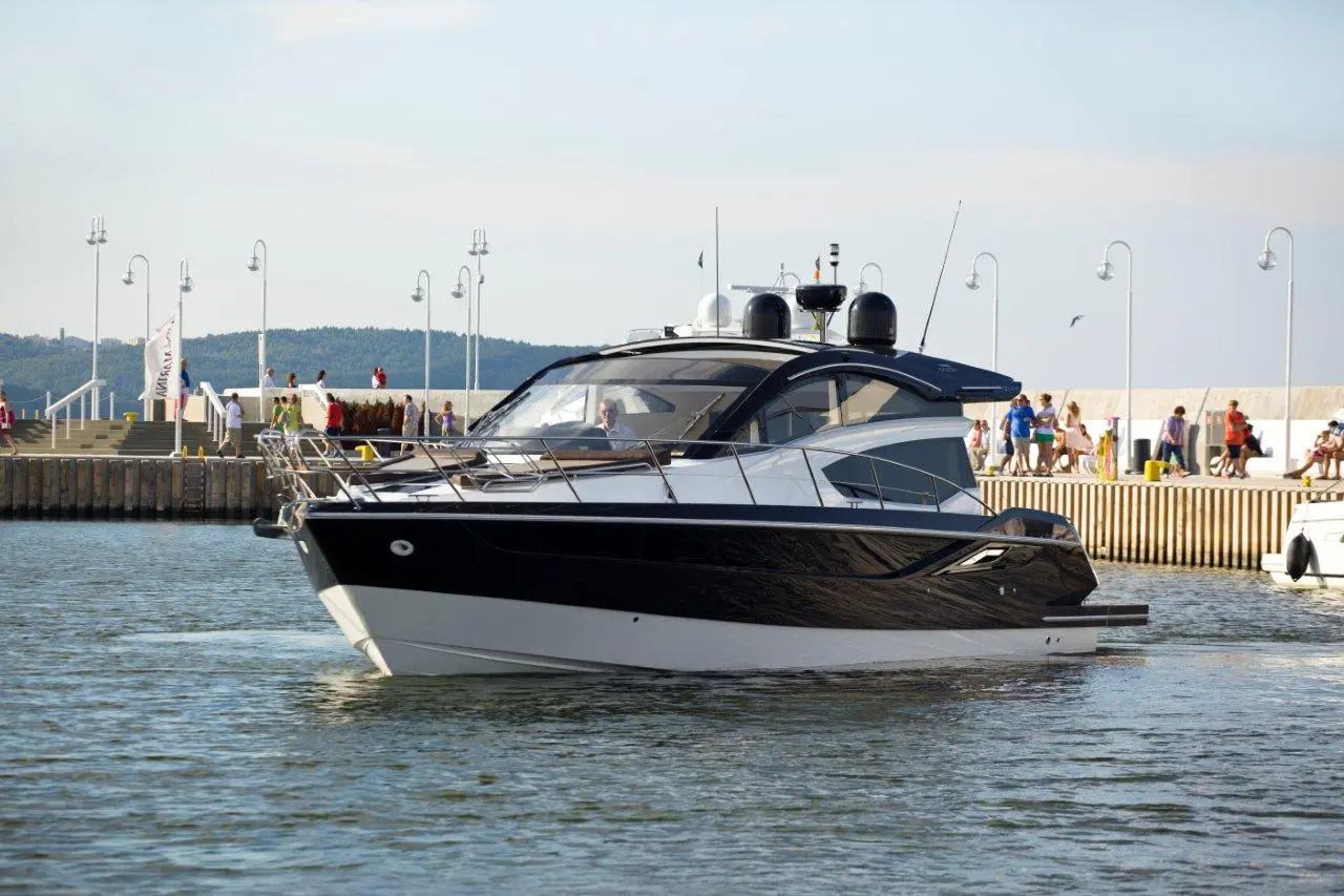 A sleek black and white yacht navigates a harbor with people walking along the promenade and scenic hills in the background.