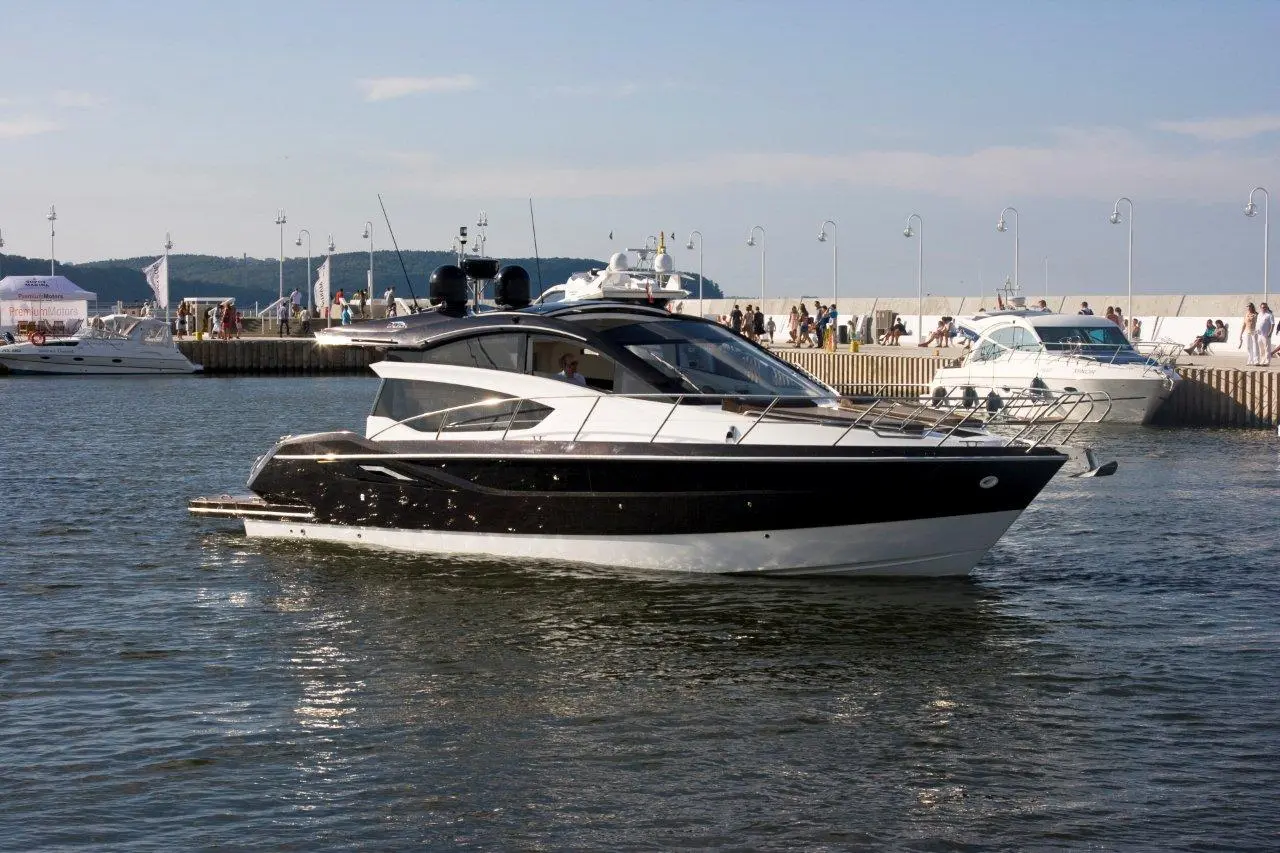 A sleek black and white motorboat navigates calm waters near a bustling pier with people and other boats in the background.