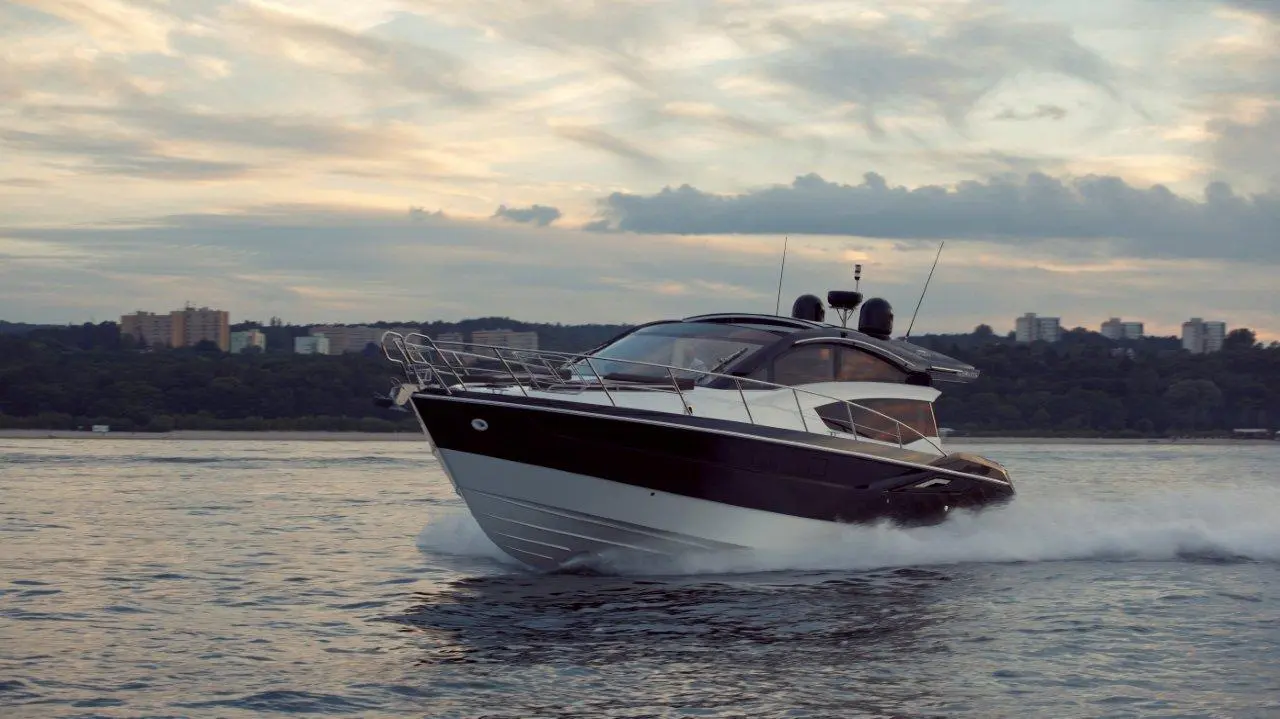 A sleek motorboat speeds through water, leaving a wake, with a city skyline and cloudy sky in the background.