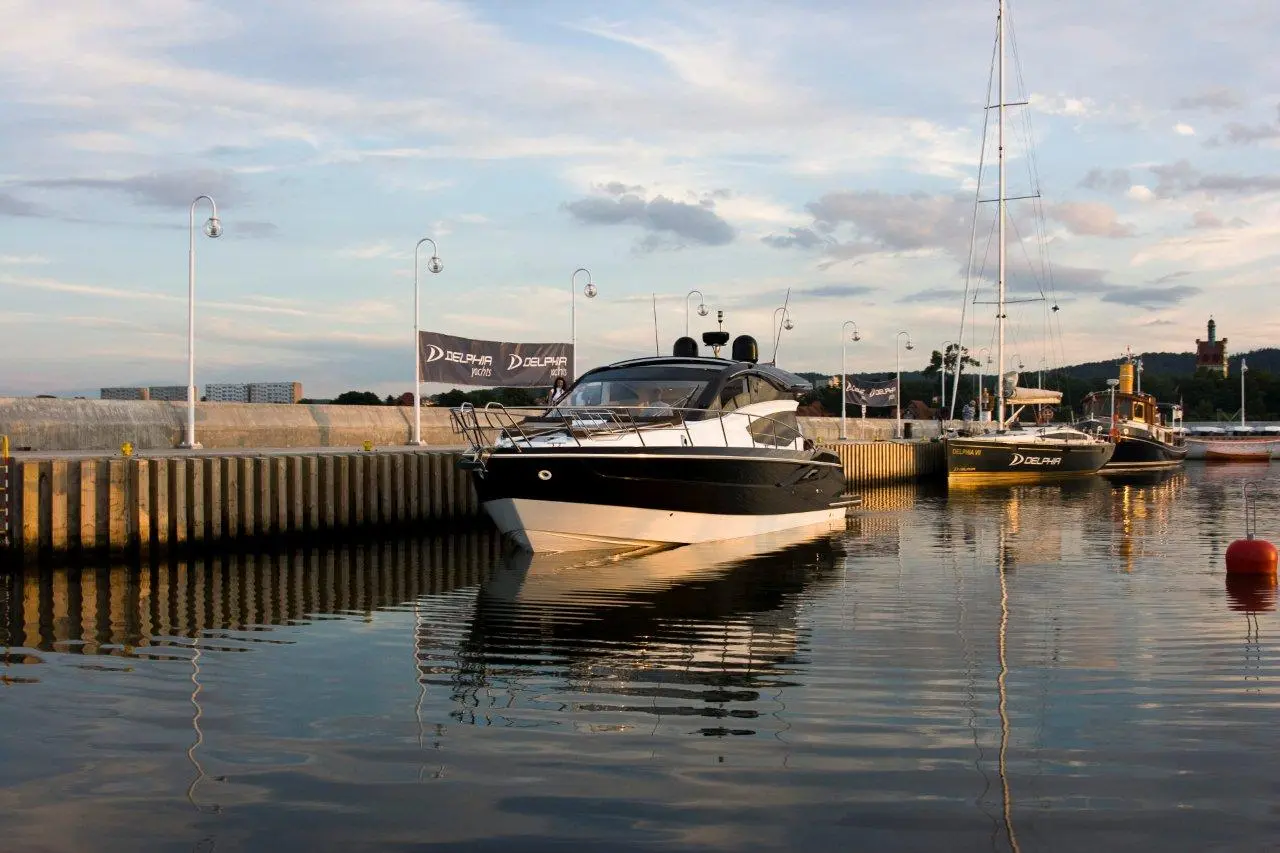 A black and white boat docked in a calm harbour, with reflective water and DELPHIA flags in the background.