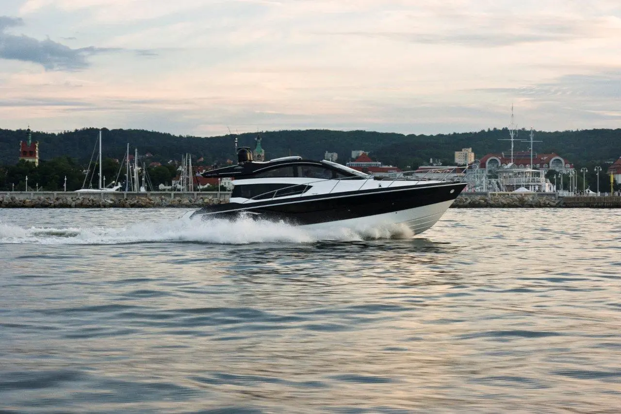 A sleek black and white speedboat cruises over the water near a harbor with buildings and masts in the background.