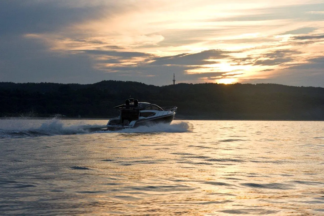 A sleek motorboat creates a wake on calm waters during sunset, with trees and a communication tower silhouetted in the background.