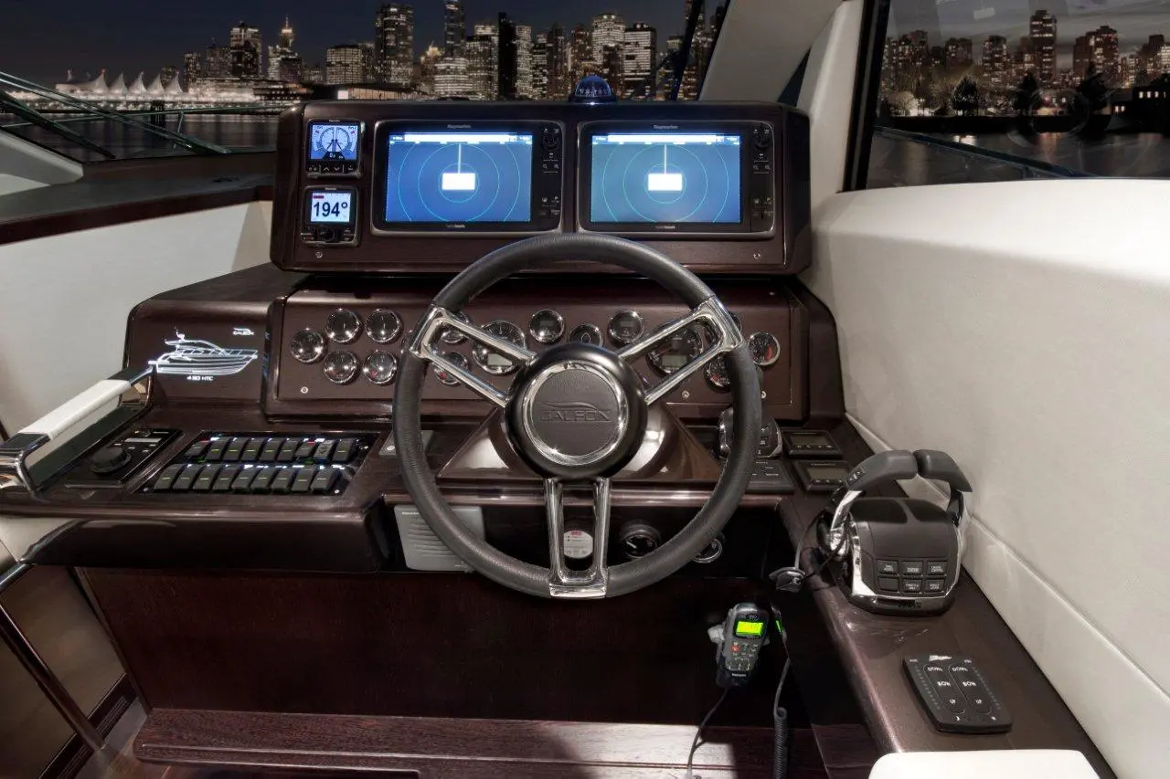 Luxury yacht control panel showcasing steering wheel, marine monitors, navigation controls, and city skyline in background.