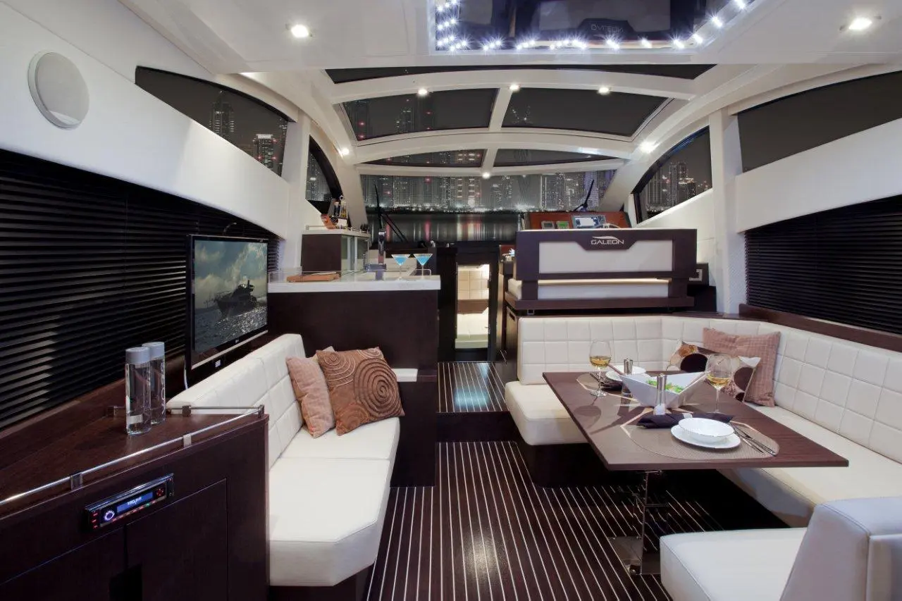 Interior of a modern yacht featuring a dining area, plush seating, a television, and city lights visible through large windows.