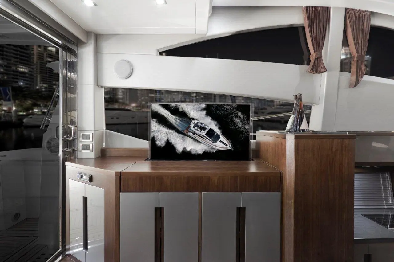 A luxury yacht interior features a sleek wooden cabinet with a TV displaying a speedboat cutting through water, with city lights visible outside.
