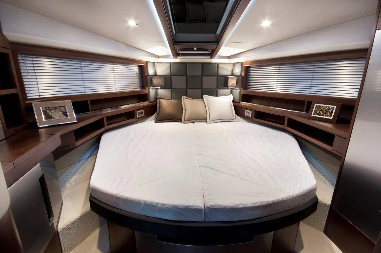 Modern yacht cabin featuring a round bed, brown and white pillows, wooden shelves, and blinds on the windows.