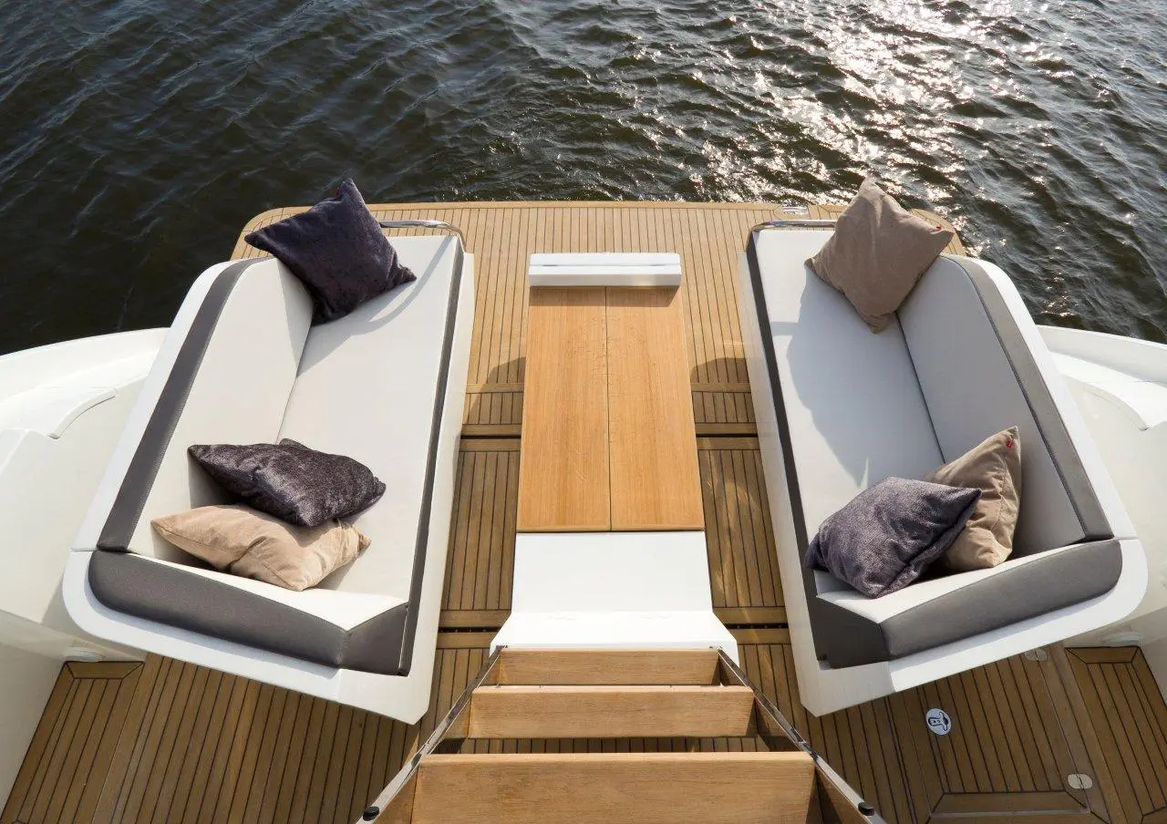 A stylish boating deck features two grey cushioned sofas, a wooden table, and steps leading downwards, overlooking water.