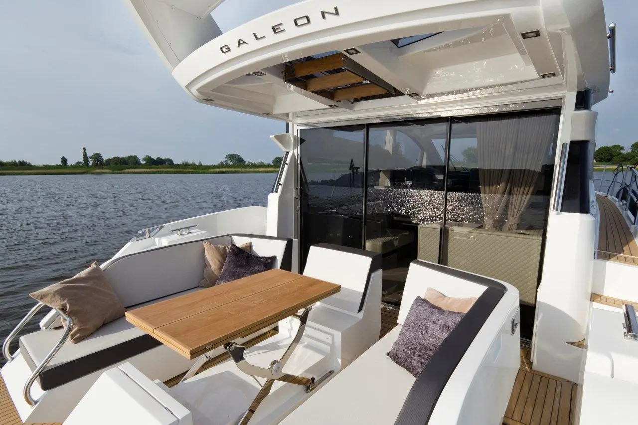 Luxury yacht deck featuring a wooden table, plush cushions, and panoramic views of the water and greenery. Brand: Galeon.