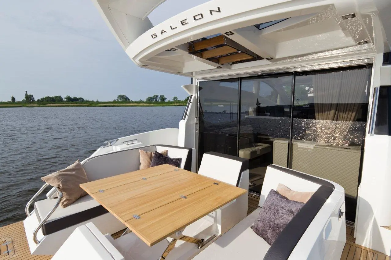 Modern boat deck featuring a wooden table, white chairs, and cushions, overlooking calm waters and green shores.