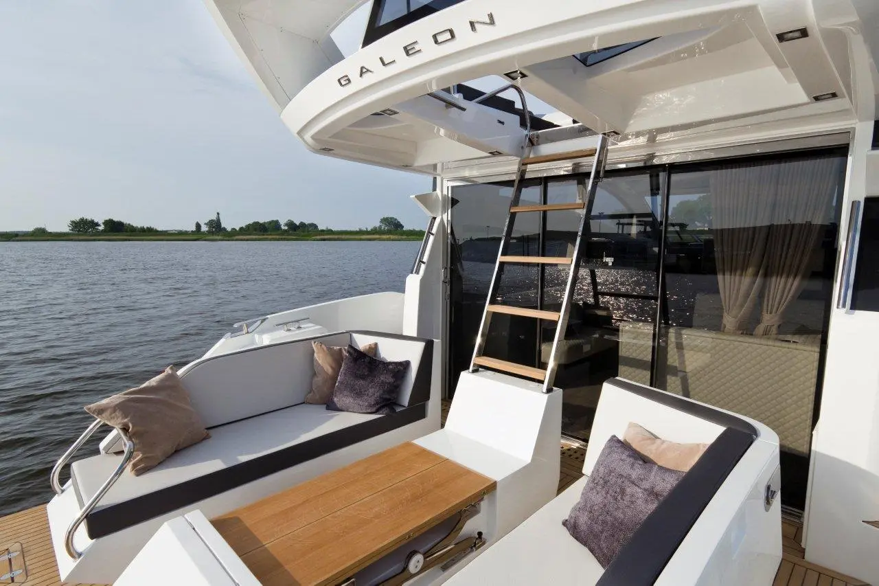 Luxury boat deck featuring a wooden table, gray seating with cushions, and a retractable ladder. Galeon branding visible.
