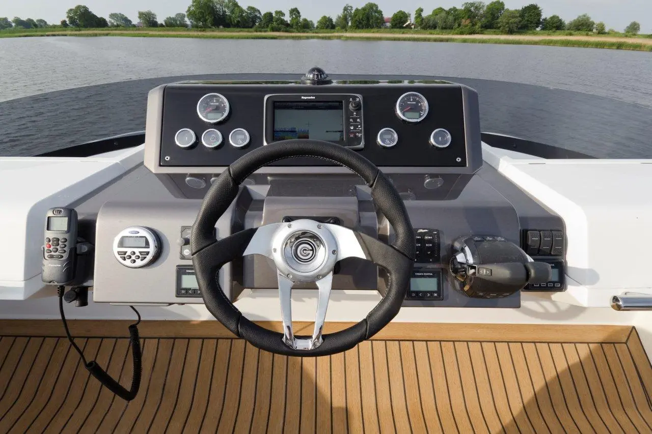 Boat steering wheel and dashboard with gauges, controls, and a view of water and trees in the background.