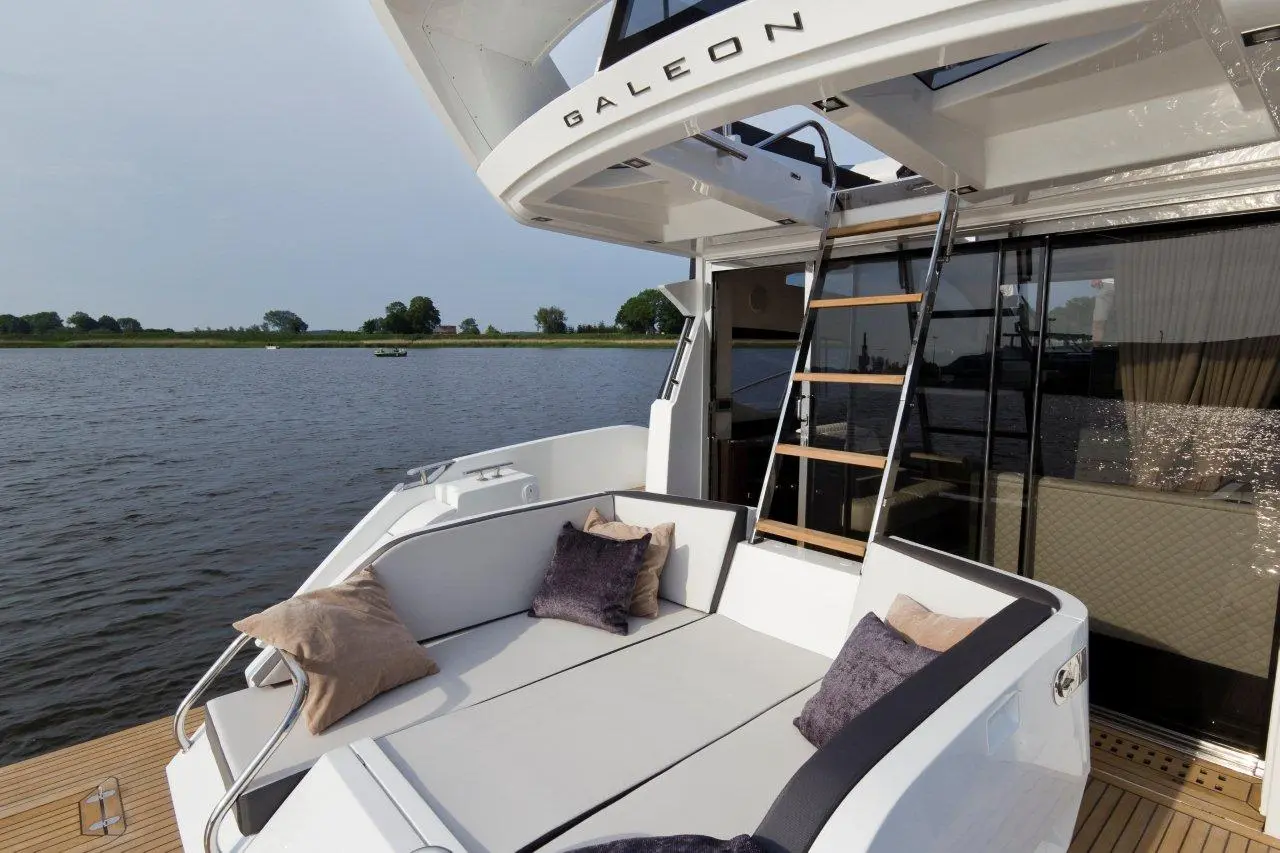 Luxury yacht seating area with pillows, stainless steel railings, and ladder; scenic waterfront in the background.