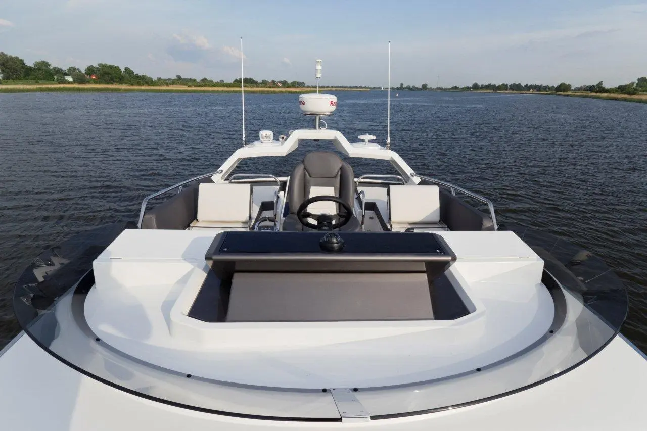 View from the bow of a sleek motorboat with a steering wheel and seating area, surrounded by calm water and greenery.