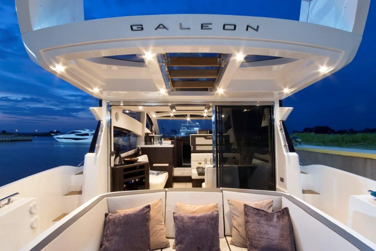 Luxury yacht interior view of a Galeon model, featuring modern furniture, lighting, and plush cushions against a twilight backdrop.