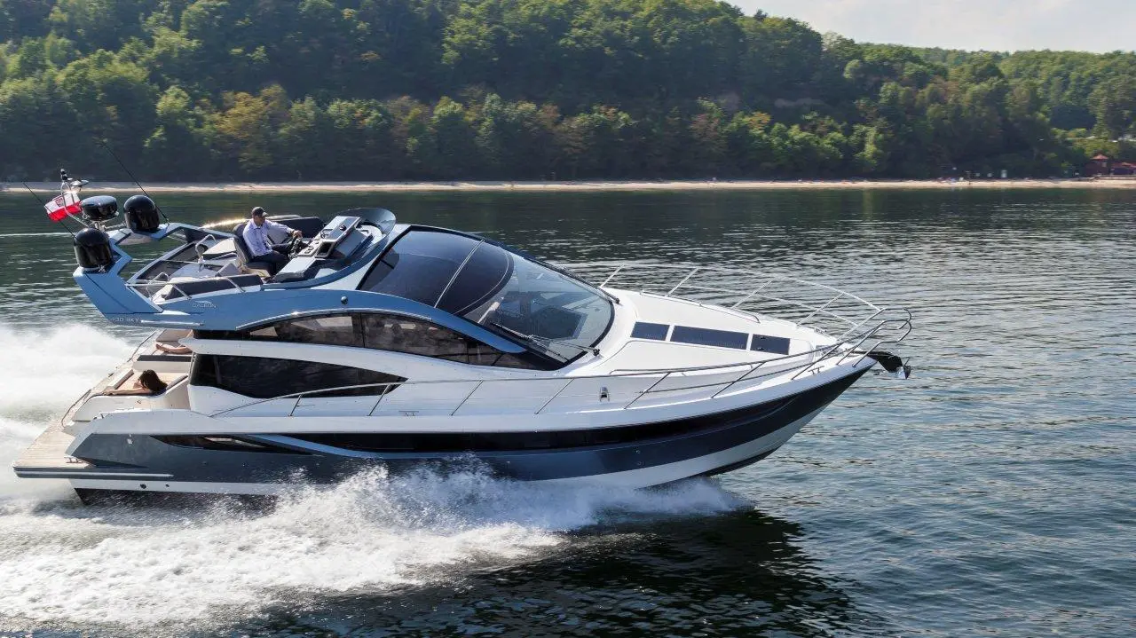 A sleek motorboat speeds across calm water, with a man at the helm and lush green trees in the background.