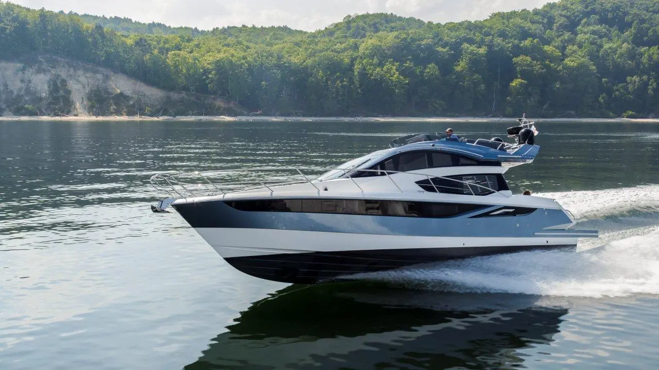 A sleek motor yacht speeds across a calm lake, surrounded by green hills and trees, with people enjoying the ride.