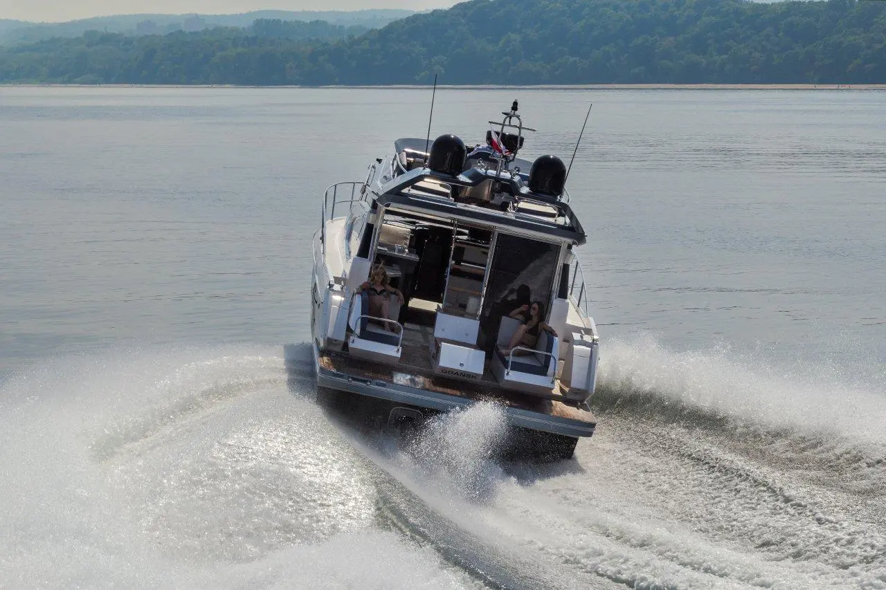 A luxury motor yacht speeds through calm water, leaving white wake; two passengers relax in deck chairs at the rear.
