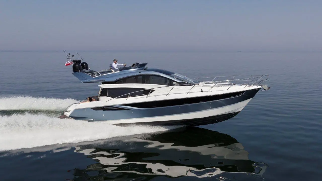 A sleek motor yacht gliding across calm waters, with a man steering at the helm under clear skies.
