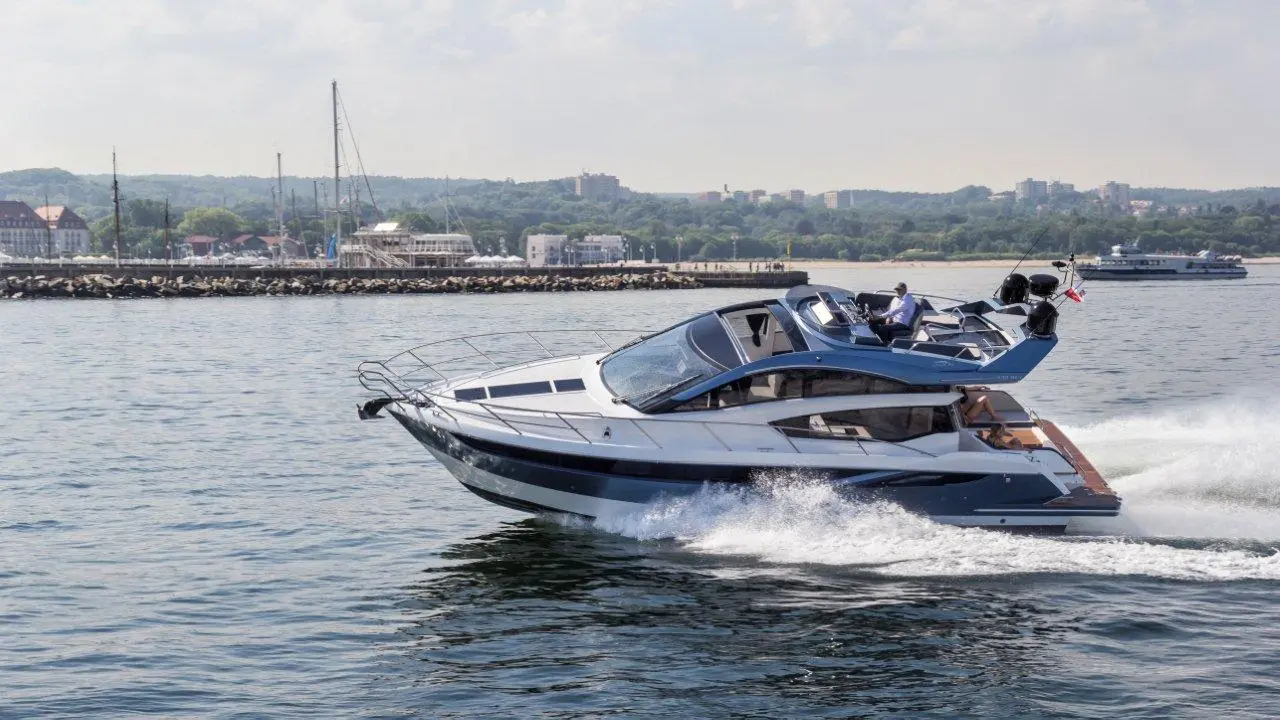 A modern motorboat speeds across a calm water surface near a harbor with sailboats and buildings in the background.