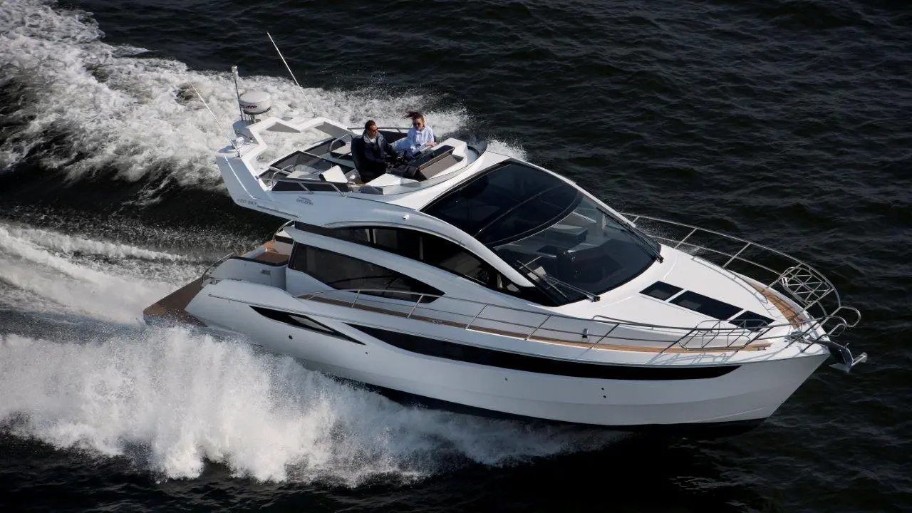 Luxury motor yacht cruising on water, with two individuals at the helm, surrounded by white wake and a clear blue background.