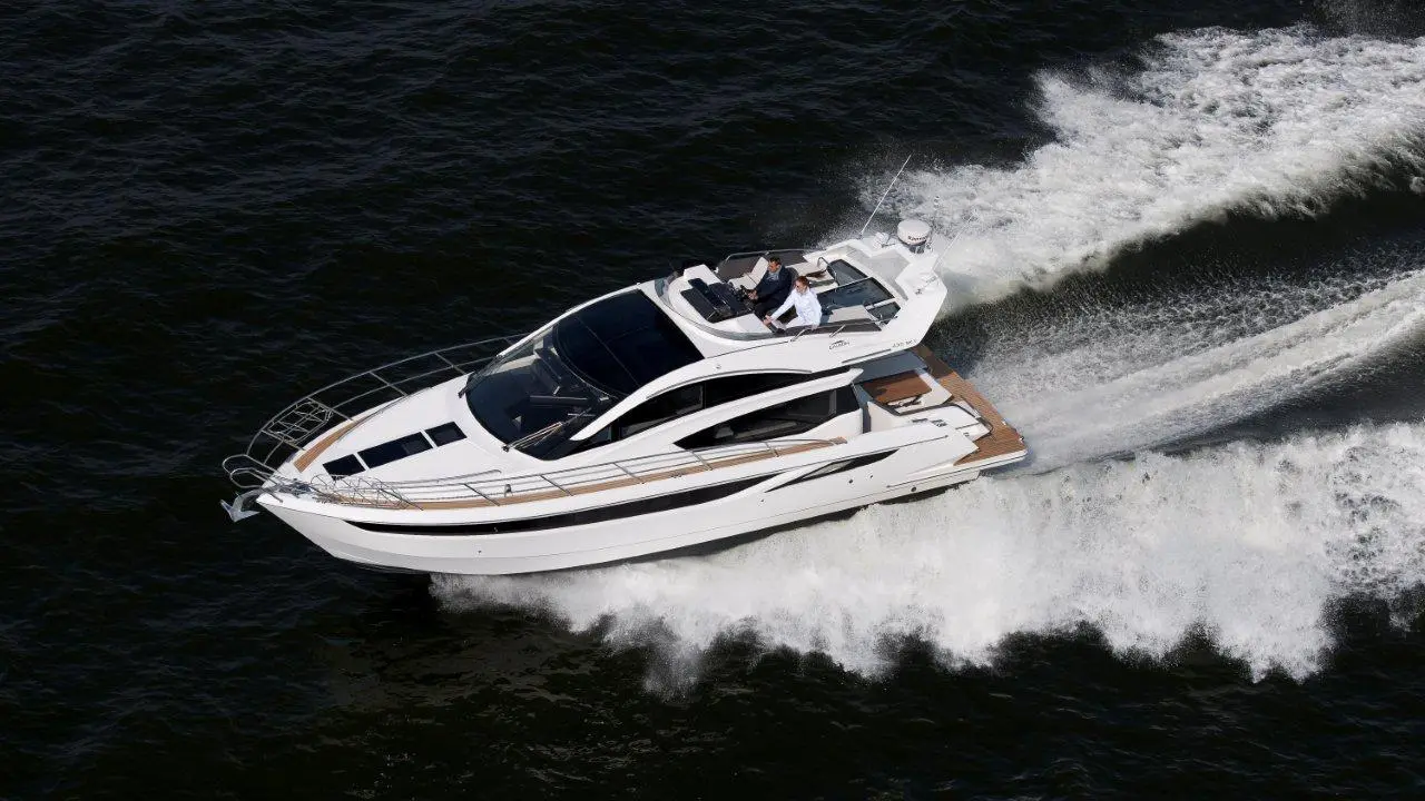 A sleek white yacht speeds over dark waters, creating frothy waves; two people are onboard, navigating the vessel.