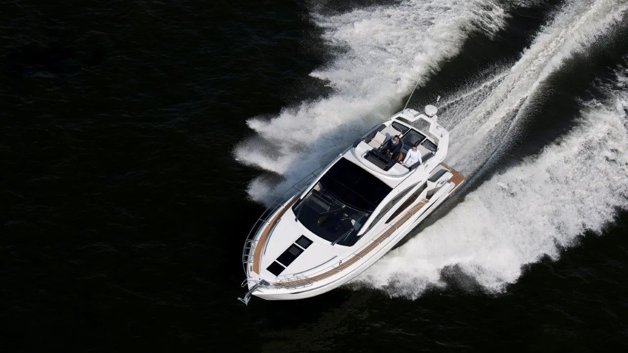 A sleek white motor yacht speeds through dark water, leaving a trail of white foam, with two people on board enjoying the ride.