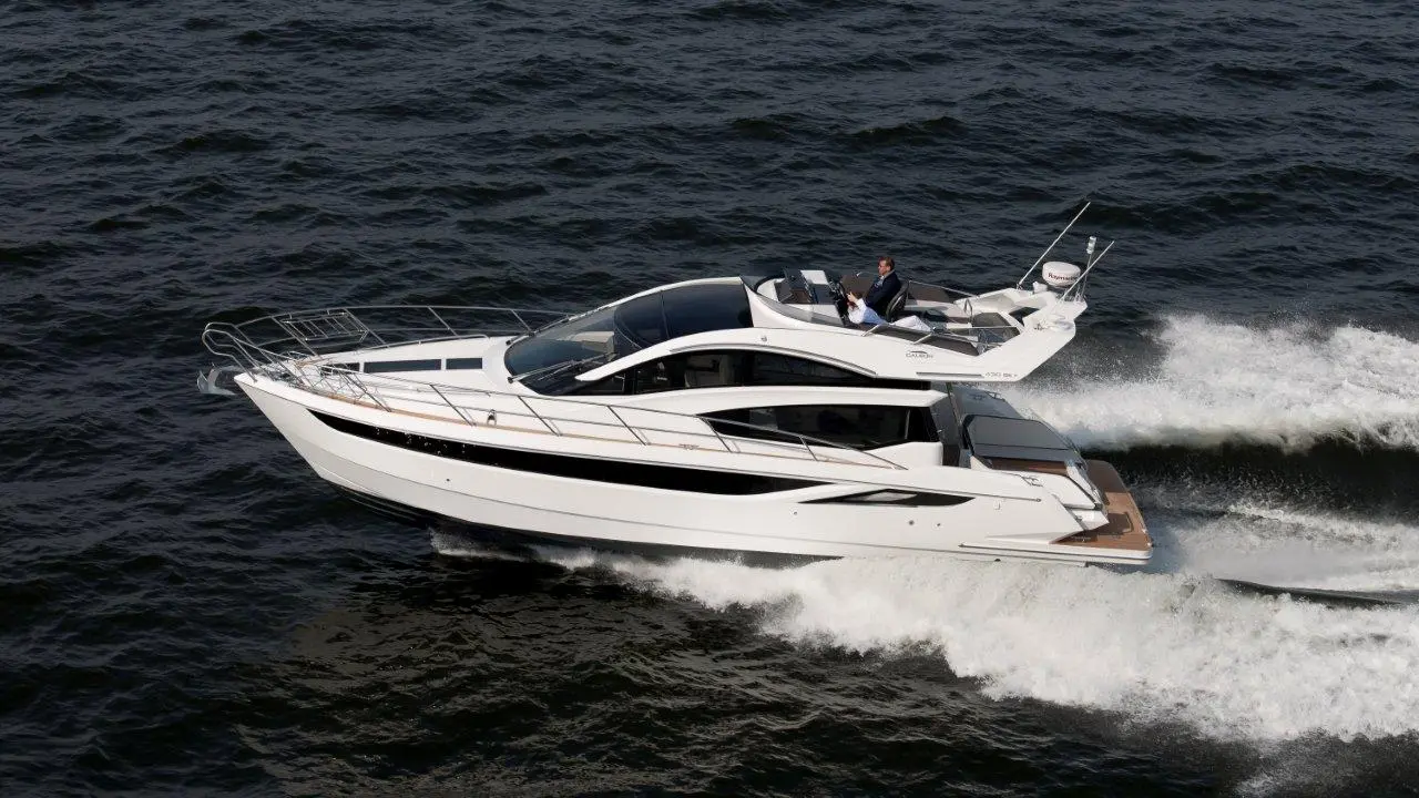 A sleek white motorboat cruises through dark water, with a man steering from the top deck under bright sunlight.