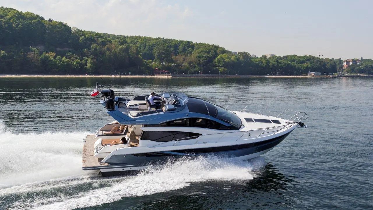 A sleek motor yacht speeds through calm waters, showcasing modern design against a green, wooded shoreline.