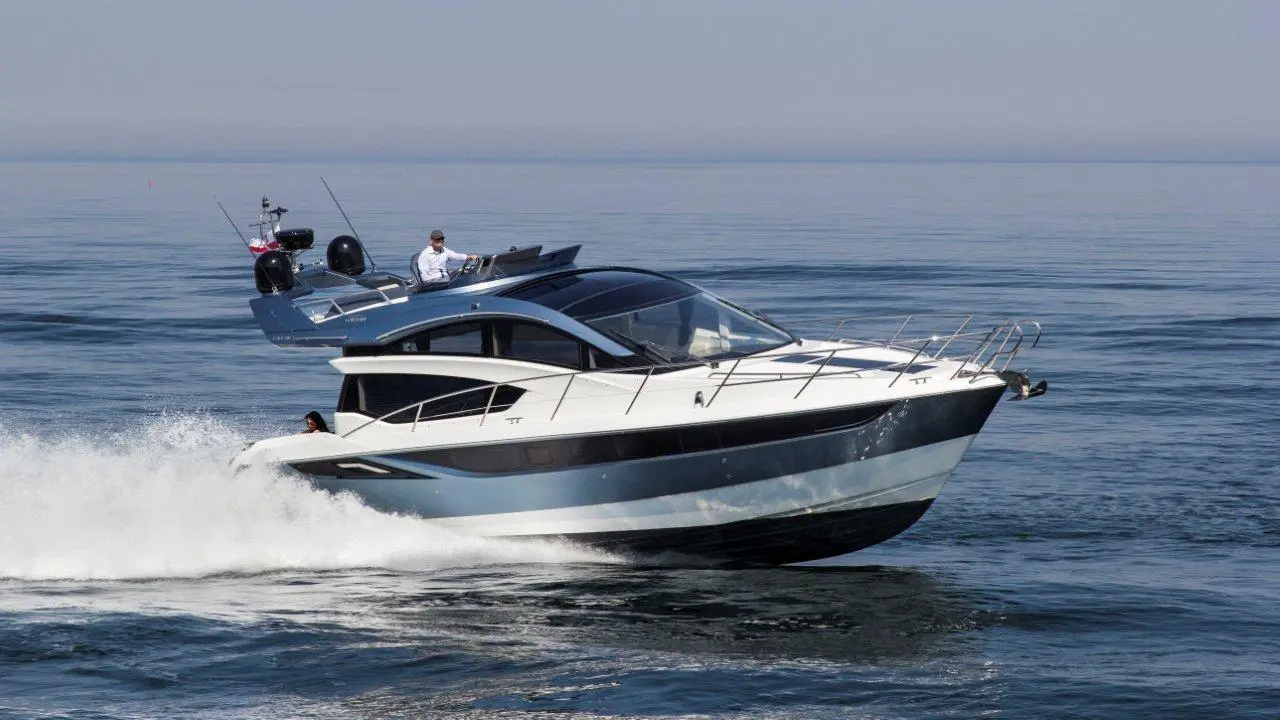A sleek powerboat glides over calm waters, creating splashes; a captain steers from the driver's seat under clear skies.