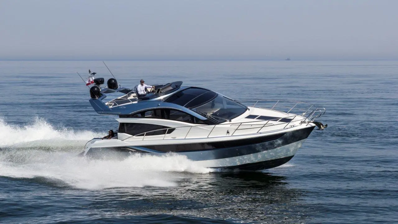 Luxury motor yacht cruising on calm waters, creating a wake; a man is steering while another person relaxes onboard.