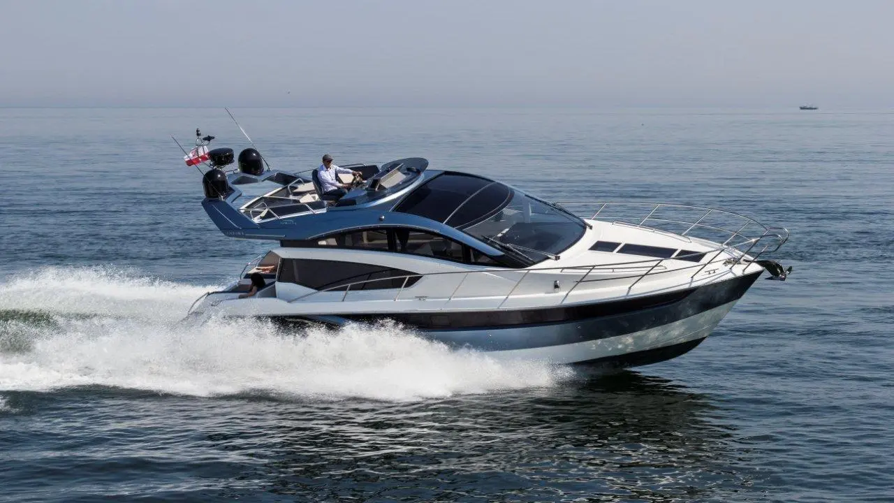A sleek motorboat cruises over calm water, with a man at the helm and an English flag flying above.