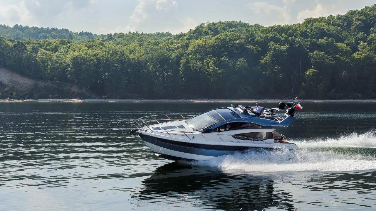 A sleek motorboat speeds across a calm lake, surrounded by lush green trees and a clear blue sky.